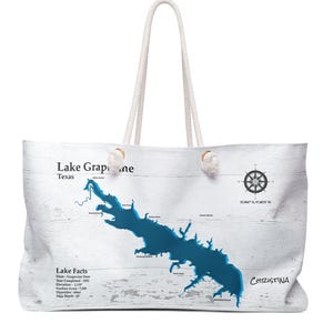 May include: White tote bag with a blue map of Lake Grapevine, Texas, and the name "Christina". The bag features a nautical compass and lake facts. It has white rope handles and is designed for carrying items.