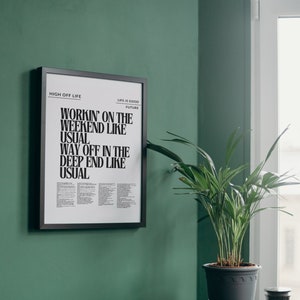 Peut inclure: Une affiche encadrée en noir avec du texte blanc qui dit "HIGH OFF LIFE", "LIFE IS GOOD FUTURE", "WORKIN' ON THE WEEKEND LIKE USUAL", "WAY OFF IN THE DEEP END LIKE USUAL". L'affiche est accrochée à un mur vert. Une plante en pot est posée sur une table d'appoint noire.