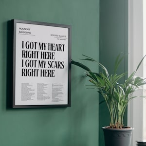 May include: A black framed print with the lyrics "I got my heart right here I got my scars right here" from the song "Wicked Games" by The Weeknd. The print is hanging on a green wall next to a potted plant.