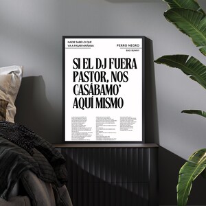 Op de afbeelding: A framed black and white poster with Spanish text, including "SI EL DJ FUERA PASTOR, NOS CASÁBAMO' AQUÍ MISMO" and "PERRO NEGRO BAD BUNNY." The poster is displayed above a black nightstand, with a plant and a wall lamp.