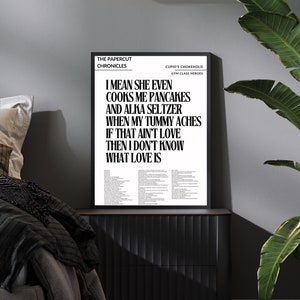 May include: Black and white typography print with the text "The Papercut Chronicles Cupid's Chokehold Gym Class Heroes I mean she even cooks me pancakes and alka seltzer when my tummy aches if that ain't love then I don't know what love is". The print is framed in a black frame and is hanging on a wall in a bedroom.