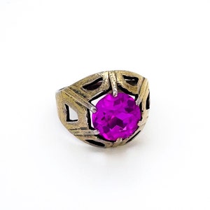 May include: A gold-toned ring with a large, round, magenta gemstone. The ring has a geometric design around the gemstone, with cut-out shapes. The ring is set against a white background.
