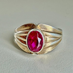 Może przedstawiać: Srebrny pierścionek z dużym, owalnym, rubinowo-czerwonym kamieniem szlachetnym. Pierścionek ma teksturowany, żebrowany wzór na obrączce, ze złotymi bokami. Kamień szlachetny jest centralnym punktem, osadzonym w srebrnej oprawie.