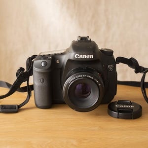 May include: A black Canon EOS 7D digital SLR camera with a lens and lens cap. The camera has a black strap and is on a wooden surface. The lens has the text "Canon Lens EF 50mm 1:1.8".