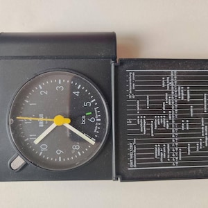 May include: Black Braun travel clock with a round face and a hinged cover. The clock face has white numerals, white hands, and a yellow second hand. The open cover reveals a world time zone chart.