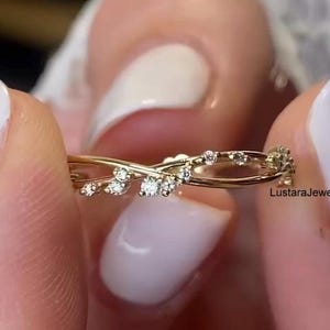 Op de afbeelding: Een delicate gouden ring met kleine, sprankelende diamanten. De ring heeft een verweven ontwerp, met de diamanten langs de rondingen gezet. De ring wordt tussen twee vingers met witte nagellak gehouden. De tekst "LustaraJewellers" is zichtbaar.