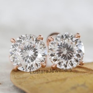 May include: A pair of round, clear diamond stud earrings with rose gold settings. The earrings are set against a blurred background with a brown leaf. The text "LustaraJewellers" is visible.