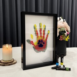May include: A framed handprint art piece with a paw print in the center. The handprint is in yellow, red, and gray paint. A lit candle and acrylic paints are also visible.