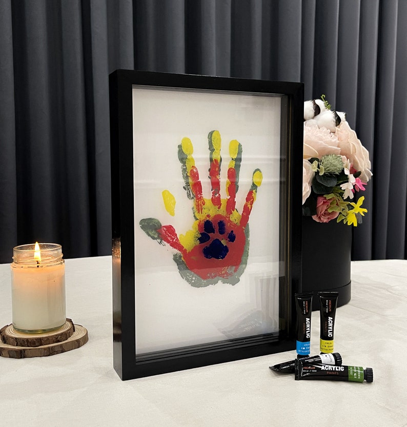 May include: A framed handprint art piece with a paw print in the center. The handprint is in yellow, red, and gray paint. A lit candle and acrylic paints are also visible.