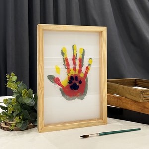 May include: A framed handprint art piece. The handprint is made with red, yellow, and gray paint, with a blue paw print in the center. The frame is made of light-colored wood.