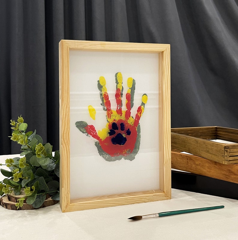 May include: A framed handprint art piece. The handprint is made with red, yellow, and gray paint, with a blue paw print in the center. The frame is made of light-colored wood.