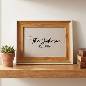 May include: A rectangular wooden picture frame with a light brown grain finish containing a white insert. The text "The Johnson" is printed in black script above the text "Est 2026" in a simple black font. The frame is positioned on a shelf.