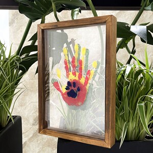 May include: A framed handprint and paw print art piece. The handprint is in red, yellow, and green paint, with a blue paw print in the center. The frame is made of brown wood and has a glass front.