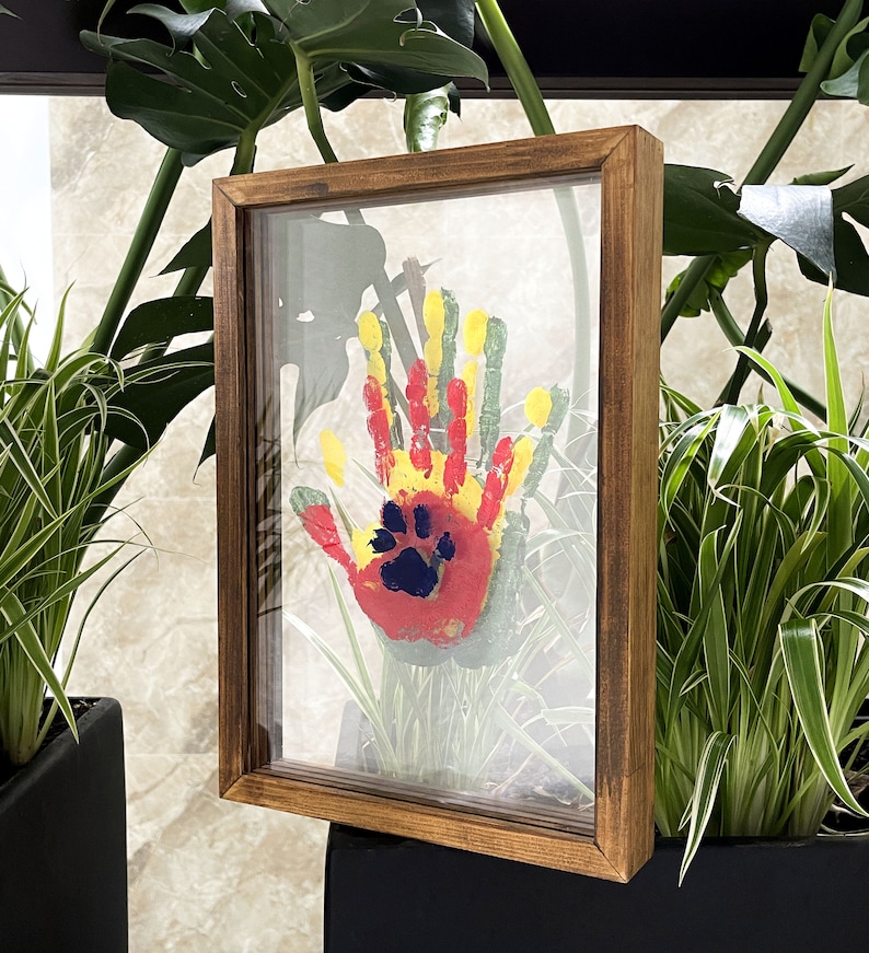 May include: A framed handprint and paw print art piece. The handprint is in red, yellow, and green paint, with a blue paw print in the center. The frame is made of brown wood and has a glass front.