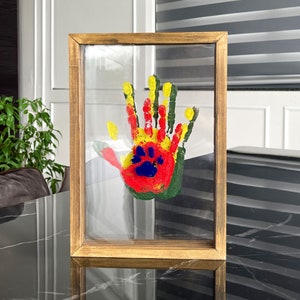 May include: A framed handprint art piece. The handprint is made with red, yellow, green, and blue paint. The frame is made of wood with a light brown finish. The artwork is displayed on a clear glass surface.