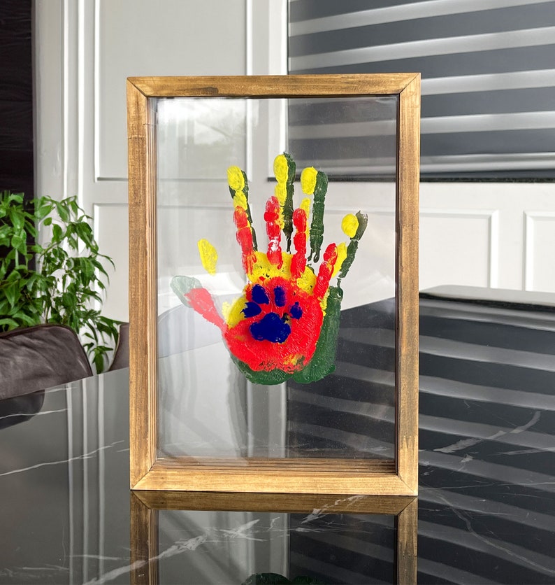 May include: A framed handprint art piece. The handprint is made with red, yellow, green, and blue paint. The frame is made of wood with a light brown finish. The artwork is displayed on a clear glass surface.