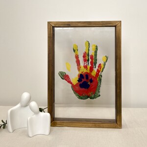 May include: A framed artwork featuring a colorful handprint with a paw print in the center. The handprint uses red, yellow, green, and blue paint. The frame is made of dark brown wood. Two white ceramic figures are in the foreground.