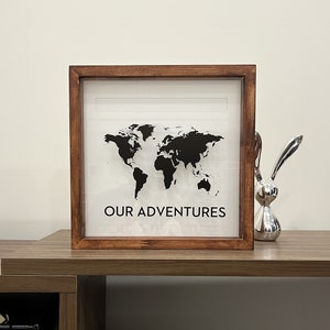 May include: A wooden frame with a world map and the text "OUR ADVENTURES" printed on a white background. The frame is sitting on a wooden surface.