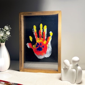 May include: A framed handprint art piece with a child's handprint and a paw print in red, yellow, and blue paint on a black background.
