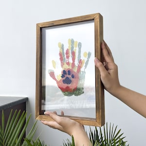May include: A framed artwork featuring colorful handprints and a paw print. The artwork is displayed in a rectangular wooden frame. The handprints are in shades of red, yellow, green, and blue. The frame is held by two hands.