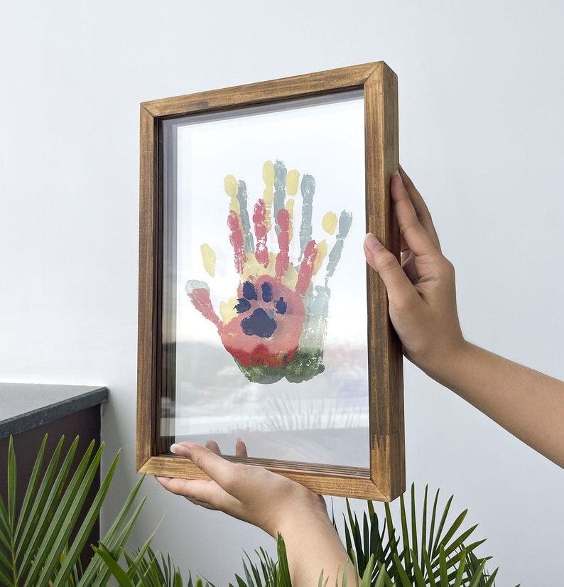 May include: A framed artwork featuring colorful handprints and a paw print. The artwork is displayed in a rectangular wooden frame. The handprints are in shades of red, yellow, green, and blue. The frame is held by two hands.