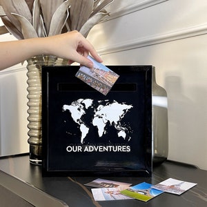 May include: Black wooden box with a world map and the text "OUR ADVENTURES" on the front. The box has a slot for inserting photos or mementos. There are several photos of travel destinations on the table in front of the box.