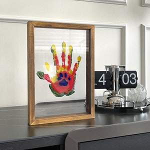 May include: A framed handprint art piece with a paw print in the center. The handprint is made with red, yellow, green, and blue paint. The frame is made of wood.