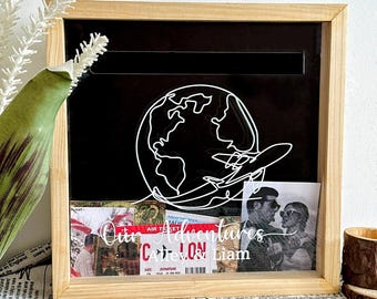 May include: A wooden shadow box with a black interior, featuring a white outline of the globe and a plane. The box contains photos and a ticket, with the text "Our Adventures Cate & Liam".