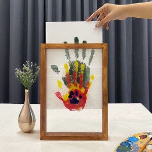 May include: A wooden framed artwork featuring a colorful handprint with red, yellow, green, and blue paint. The artwork is displayed on a white background. A hand holds a white canvas above the frame. A gold vase with white flowers is on the left.