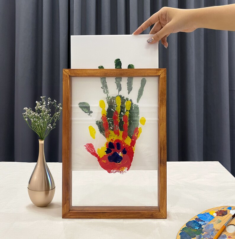 May include: A wooden framed artwork featuring a colorful handprint with red, yellow, green, and blue paint. The artwork is displayed on a white background. A hand holds a white canvas above the frame. A gold vase with white flowers is on the left.