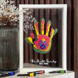 May include: A framed handprint art piece with a colorful handprint design. The handprint is painted with red, yellow, green, and blue paint, and includes a paw print. The frame is white, and the text "The Smith Family EST. 2025" is printed on the glass.