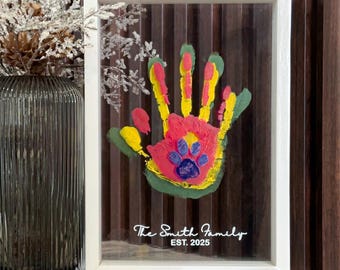 May include: A framed handprint art piece with a colorful handprint design. The handprint is painted with red, yellow, green, and blue paint, and includes a paw print. The frame is white, and the text "The Smith Family EST. 2025" is printed on the glass.