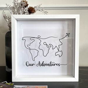 May include: White picture frame with a black line drawing of a world map and a small airplane. The text "Our Adventures" is written below the map.