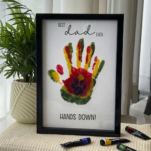 May include: Framed artwork with a black border featuring a colorful handprint in red, yellow, and green. The text "BEST dad EVER" and "HANDS DOWN!" are printed above and below the handprint. Paint tubes are visible.