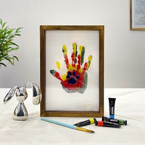 May include: A framed artwork featuring a colorful handprint with a paw print in the center. The handprint is made with red, yellow, and green paint. A silver bunny figurine, paint tubes, and a paintbrush are also in the image.
