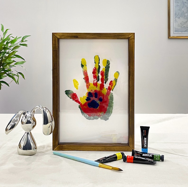 May include: A framed artwork featuring a colorful handprint with a paw print in the center. The handprint is made with red, yellow, and green paint. A silver bunny figurine, paint tubes, and a paintbrush are also in the image.