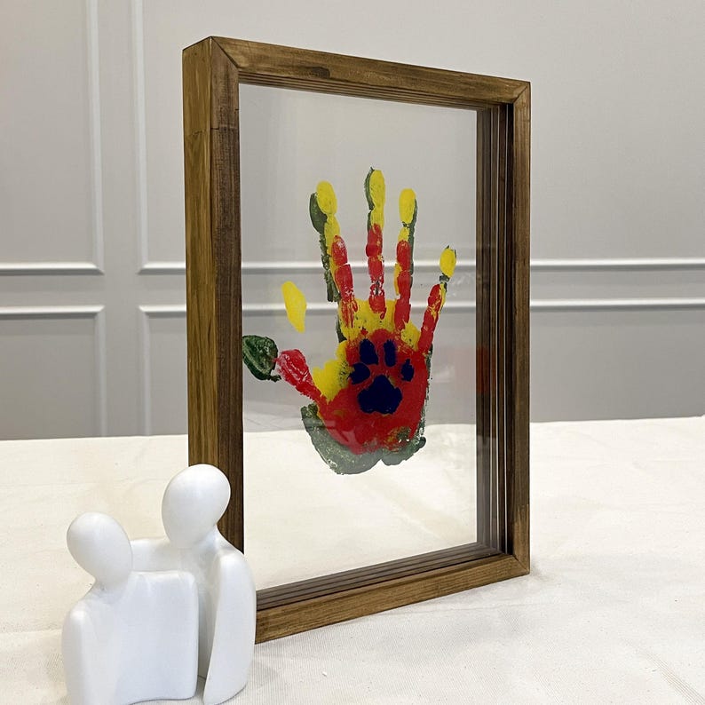 May include: A framed handprint art piece with a wooden frame. The handprint is a colorful mix of red, yellow, and green paint, with a paw print design in the center. A white sculpture of two figures is in the foreground.
