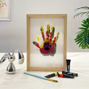 May include: A framed handprint art piece with a wooden frame. The handprint is made with red, yellow, and green paint, with a paw print in the center. A silver bunny figurine, paint tubes, and a paintbrush are also visible.