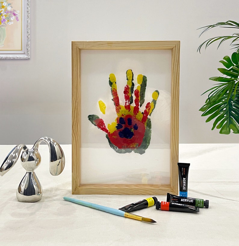 May include: A framed handprint art piece with a wooden frame. The handprint is made with red, yellow, and green paint, with a paw print in the center. A silver bunny figurine, paint tubes, and a paintbrush are also visible.
