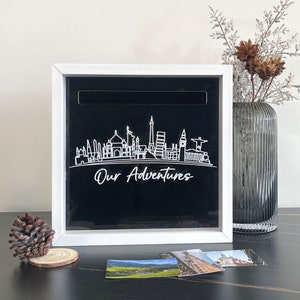 May include: White wooden box with a black background and a white outline of a city skyline. The text "Our Adventures" is written below the skyline. The box has a slot on the top for inserting items.
