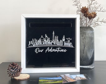 May include: White wooden box with a black background and a white outline of a city skyline. The text "Our Adventures" is written below the skyline. The box has a slot on the top for inserting items.