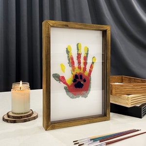 May include: A framed handprint art piece with a paw print in the center. The handprint is in red, yellow, and green paint. The frame is made of wood with a warm, brown finish. Paintbrushes and a lit candle are also in the image.