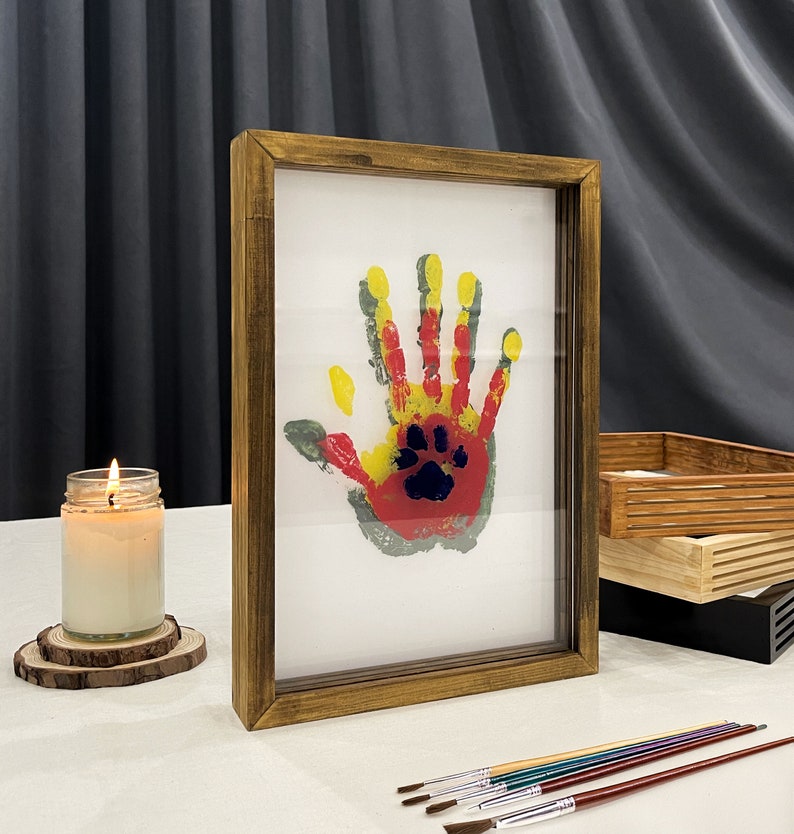 May include: A framed handprint art piece with a paw print in the center. The handprint is in red, yellow, and green paint. The frame is made of wood with a warm, brown finish. Paintbrushes and a lit candle are also in the image.