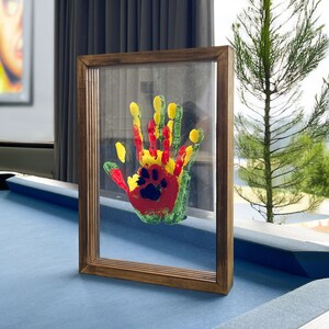 May include: A framed artwork featuring a colorful handprint with a paw print design. The handprint is painted in red, yellow, and green, set against a clear background within a brown wooden frame. The artwork is displayed on a blue surface.