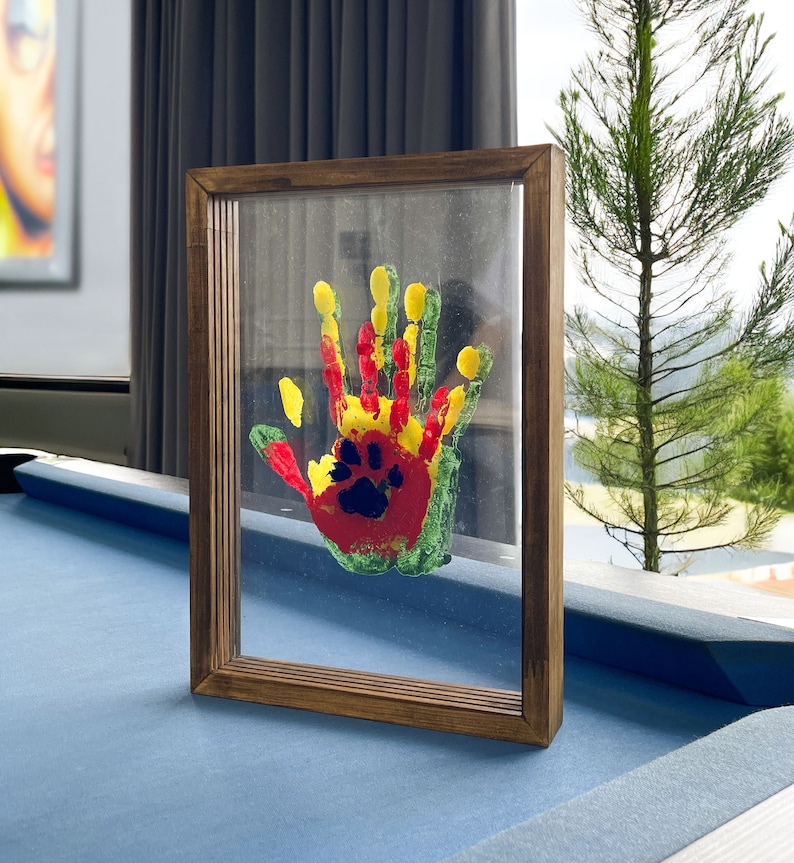 May include: A framed artwork featuring a colorful handprint with a paw print design. The handprint is painted in red, yellow, and green, set against a clear background within a brown wooden frame. The artwork is displayed on a blue surface.
