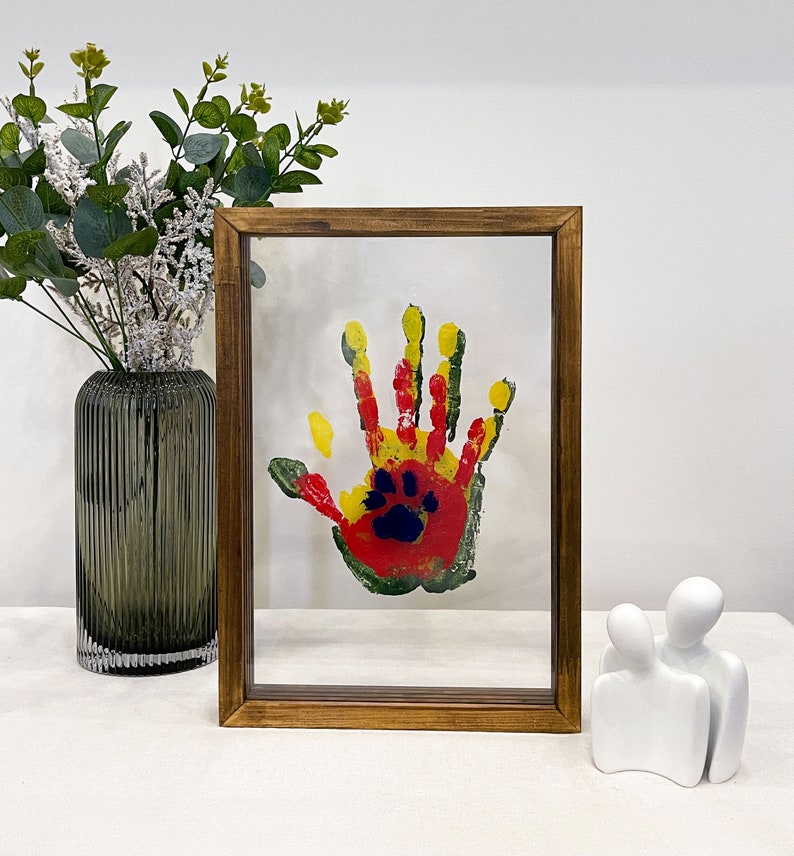 May include: A framed handprint art piece with a wooden frame. The handprint is a colorful mix of red, yellow, and green paint, with a paw print in the center. The artwork is displayed on a white background.