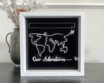 May include: White shadow box with a black background. The box contains a white wire outline of a world map with a small airplane at the top right corner. The text "Our Adventures" is written in white cursive below the map.