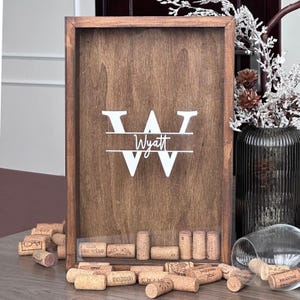 May include: A wooden shadow box with a dark brown finish, displaying the name "Wyatt" in white script within a stylized "W". The box is designed to hold wine corks, with several corks visible in the bottom.