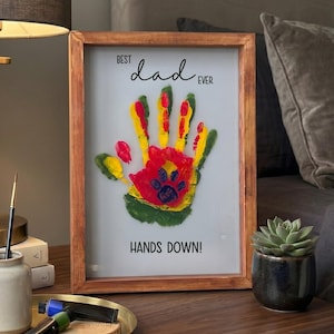May include: A framed artwork with a handprint design in red, yellow, and green, with a paw print in the center. The text reads "Best Dad Ever" and "Hands Down!" The frame is made of wood.