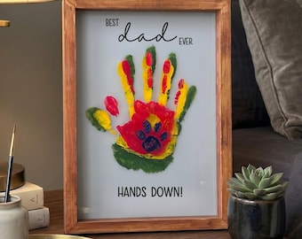 May include: A framed artwork with a handprint design in red, yellow, and green, with a paw print in the center. The text reads "Best Dad Ever" and "Hands Down!" The frame is made of wood.